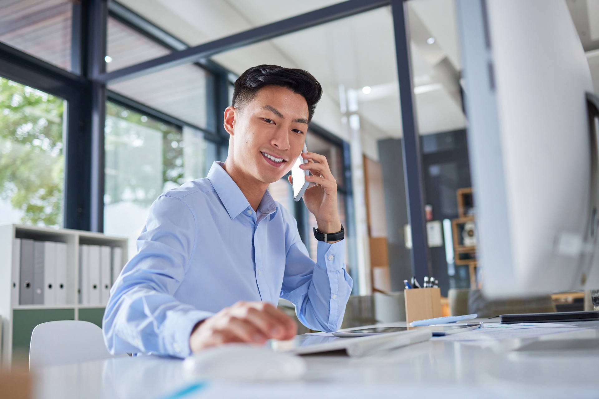 Young asian businessman making a call on his cellphone. Happy businessman typing on his computer. Business professional working in his office. Businessman calling on his smartphone.